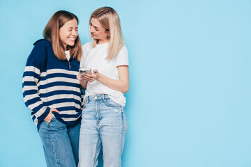 Naklejka premium Two young beautiful smiling brunette hipster female in trendy summer clothes. Carefree women posing near blue wall. Positive models having fun. Cheerful and happy. Holding smartphone. Using apps