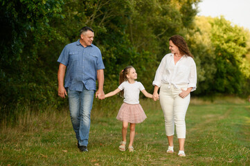 Fototapeta premium Family spending time together in summer. Mom, dad, girl walking along park path among trees, holding hands, smiling happily, looking at each other. Family, love and parenthood concept, happy childhood