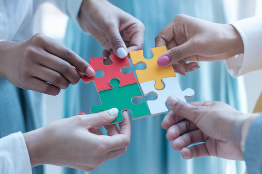 Diverse hands connecting colorful puzzle pieces, symbolizing teamwork and problem-solving. Highlights skin tone diversity and collaboration against a softly blurred neutral background
