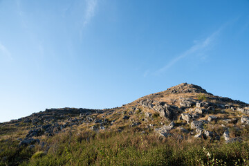 A rocky hill rises under a clear blue sky, surrounded by lush greenery, showcasing a serene and natural landscape.