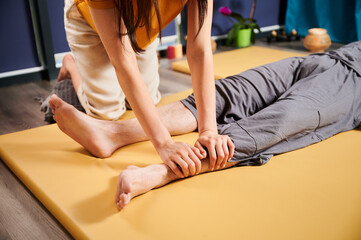 Fototapeta premium Masseuse making thai yoga massage. Cropped view of woman therapist doing traditional massage treatment for male patient. Client lying on mat, meditating, enjoying hand massage in calm atmosphere.