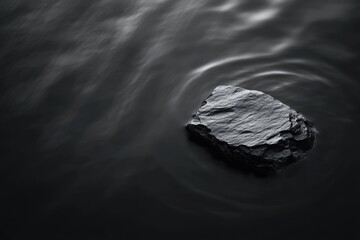 Naklejka premium Dark, solitary rock rests on calm, black water, creating ripples.