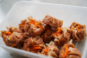 A close-up shot of several steamed pork dumplings with carrots in a clear plastic container. A fork is used to take one. Delicious Steamed Chikcen Dumplings in a Plastic Container chicken siu mai