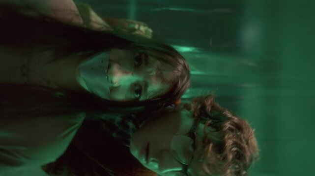 Evil maniac standing behind frightened hostage with taped mouth, both looking into camera in dimly lit basement with green illumination. Rolling camera shot, video portrait