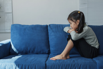 Sad little girl sitting alone on a blue sofa, arms crossed over her knees, expressing feelings of...