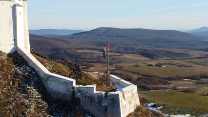 Hungarian flag waves proudly atop the fortress bastion, surrounded by rolling hills and a picturesque valley stretching beneath a blue sky. Fuzer castle wall, with its white stone battlements.