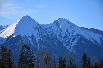 Fototapeta premium Majestic snow-covered High Tatras peaks rise dramatically against the blue sky, creating a breathtaking and serene landscape. Sunlight, rugged rock formations, slopes and summit. 