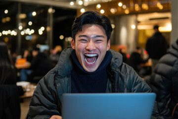 Obraz premium Young asian male laughing enthusiastically while using laptop in cozy cafe Laugh at Work Week
