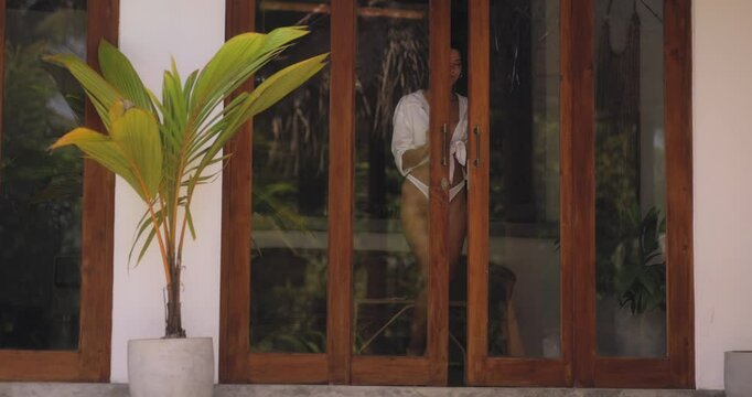 Confident woman in a white bikini with a tied shirt, standing in the doorway of a tropical villa, basking in the warm ambiance. Ideal for travel, summer fashion, and lifestyle themes.