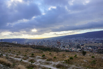 Western part of Tbilisi with modern buildings and residential houses. TV tower is visible. Cloudy sky and mountains.