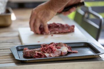 Homme découpant un morceaux de viande de sanglier. Sa planche à découper est blanche. Il est en extérieur, sur une table en bois, un plateau à ses côtés.