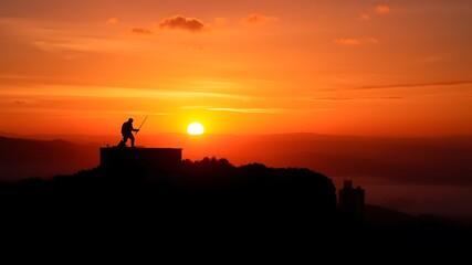 Naklejka premium A lone hiker silhouetted against a vibrant sunset, standing on a mountaintop structure, overlooking a misty valley with distant buildings, capturing the breathtaking view.