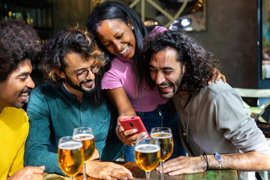Group of cheerful multi-ethnic friends enjoying drinks at a bar, laughing together while looking at a smartphone