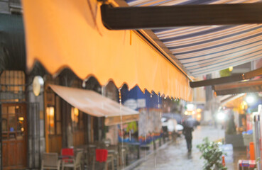 Naklejka premium Rain falls on vibrant street cafes with colorful awnings