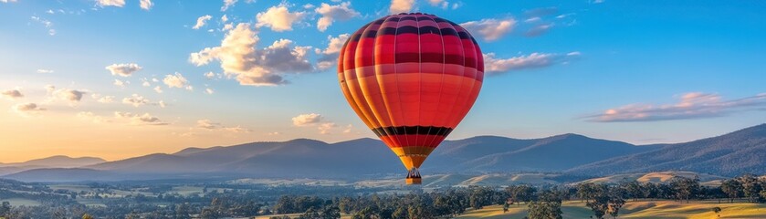 Naklejka premium A vibrant hot air balloon floats over a picturesque landscape during sunset, with mountains in the background and a colorful sky.