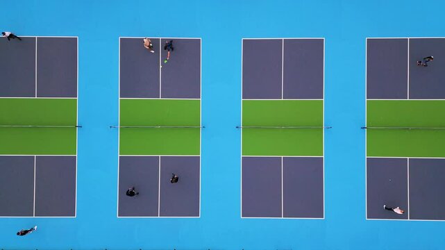 Aerial top-down view of multiple pickleball courts with players engaged in a match. The vibrant blue and green surfaces create a striking geometric pattern, capturing the dynamic movement and energy