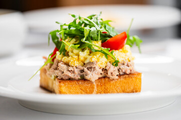 Delicious open-faced sandwich featuring a layer of creamy tuna mixed with herbs, topped with fluffy scrambled eggs, fresh arugula, and a slice of tomato, all served on crusty toasted bread.
