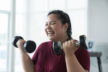 Happy smiling Asian young plus size woman lifting dumbbell weight in fitness club. Young overweight...