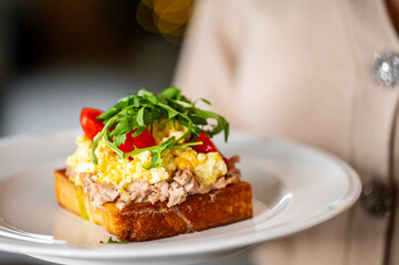 A delicious open-faced sandwich featuring toasted bread topped with tuna salad, fluffy scrambled eggs, cherry tomatoes, and fresh arugula, beautifully arranged on a white plate.