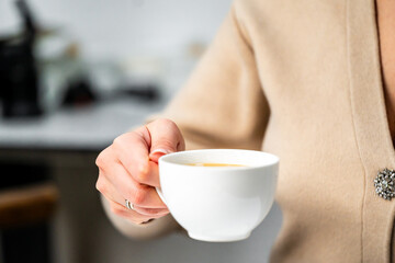 A close-up of a hand holding a white coffee cup, featuring warm beverage inside. The background is softly blurred, creating a cozy atmosphere suitable for cafe or home settings.