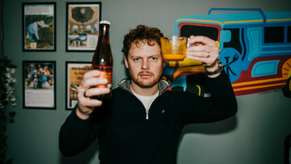 A man is holding a bottle of beer and a glass of orange juice. He is smiling and he is enjoying his drink
