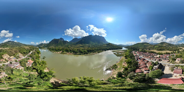 Aerial view of serene village by the river surrounded by lush mountains and bright sky, Pak Ou District, Laos.