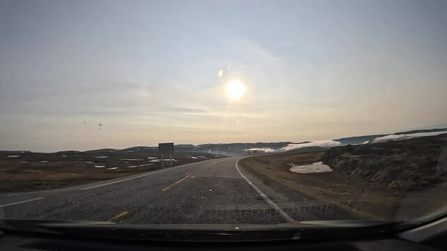 Driving through the tundra in northern Norway on the E69 road. The landscape features plains and hills covered with moss and lichen, with patches of snow. The sun shines directly ahead.
