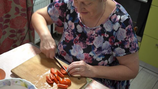An elderly woman cuts tomatoes and prepares a salad at home in the kitchen. Old hands and knife close up. Wooden board. Food vegetables. Healthy eating