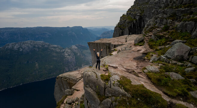 Aerial view of a breathtaking cliff and mountain landscape with a woman hiking overlooking a serene fjord, Preikestolen, Norway.