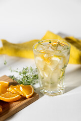 ice tea with citrus fruits and thyme in a glass on white background. iced tea with lemon and orange in drinking glass with fruits on cutting board with towel on side. harsh lighting with shadows.