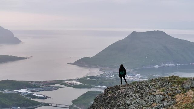 Aerial view of a serene woman standing on a rugged rock overlooking Captains Bay with majestic mountains and a calm ocean, Unalaska, United States.