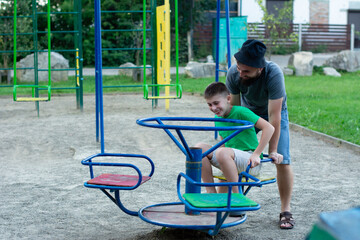 A father gently assists his son as they spin on a small merry-go-round at a playground. The scene...