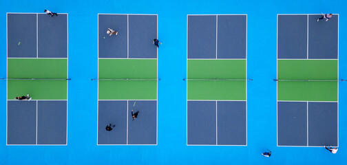 Aerial top-down view of multiple pickleball courts with players engaged in a match. The vibrant blue and green surfaces create a striking geometric pattern, capturing the dynamic movement and energy