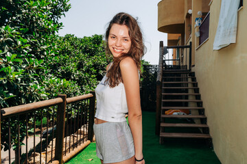  Smiling young woman leans on a wooden railing surrounded by lush greenery, enjoying a sunny day outdoors in casual wear