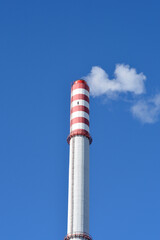 A tall industrial chimney with red and white stripes emits a wisp of white smoke against a clear blue sky