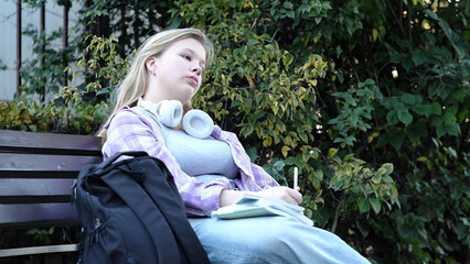 blonde girl, wearing headphones and a plaid shirt, concentrates on her studies outdoors. The image...