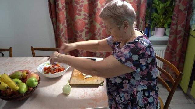 An elderly woman cuts onion and prepares a salad at home in the kitchen. Old hands and knife close up. Wooden board. Food vegetables. Healthy eating