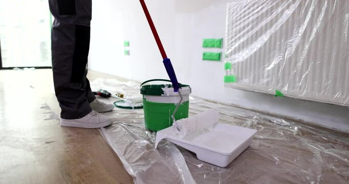 Experienced repairman dips paint roller into tray filled with white paint. Floor and furniture covered with protective foil sheets. Slow motion