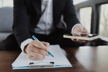Businessman in a black suit writing on a clipboard with a pen, , holding his phone and focusing on paperwork, contract signing, or business documentation, symbolizing professional work.