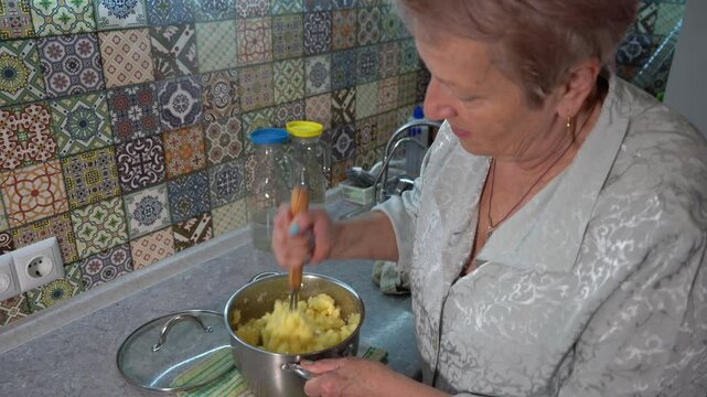 An elderly woman mashes boiled potatoes for mashed potatoes for lunch at home in the kitchen. Hands and pot close up. Food cooking table