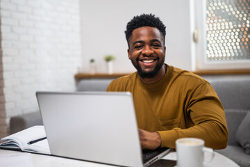 Portrait of cheerful young black businessman using laptop while enjoying  working remotely at his comfortable home.