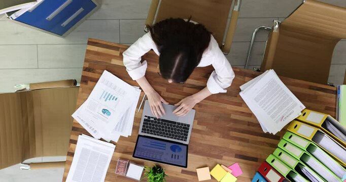 Businesswoman works on laptop with documents while colleague brings papers stack in office. Female employee feels burnout of paperwork overload at desk