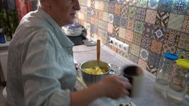 Elderly woman mashes boiled potatoes for mashed potatoes for lunch at home in the kitchen. Hands and pot close up. Food cooking table