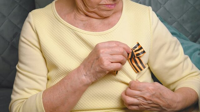 An elderly woman holds a St. George ribbon in her hands and pins it on her chest. The St. George ribbon is a symbol of May 9, the Victory Day of Russia in 1945