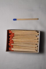 A solitary blue-tipped match next to a full box of matches with red tips, set against a neutral backdrop.