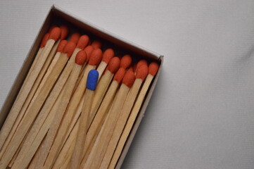 A solitary blue match contrasts vividly among a group of red matches inside a matchbox, creating a...