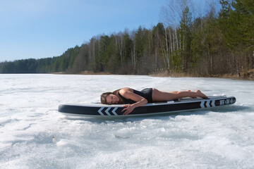 Woman in swimsuit basking in sun lying on sup board on frozen ice on river lake. Brunette female smiling posing looking at camera enjoying cold sunny day on nature, outdoor recreation on supboard.