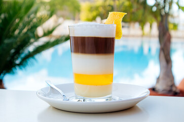 Local coffee drink in glass called The Barraquito on Tenerife island in Spain. Beautiful colorful drink against blue pool and palm trees.

