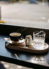 Minimalist coffee setup with a steaming espresso, a glass of water, and a milk jug on a wooden tray by the window.