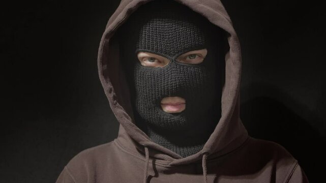 Face covered with balaclava, mask. Portrait of criminal, hacker, anonymous man in hoodie on black background.